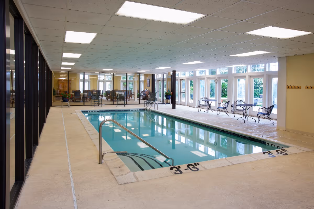 Indoor swimming pool area with clear water, metal handrails, and depth markings on the pool edge. The pool is surrounded by a beige tiled floor and large windows letting in natural light. There are tables and chairs along the far side near the windows, and a separate room with additional seating visible through glass walls.