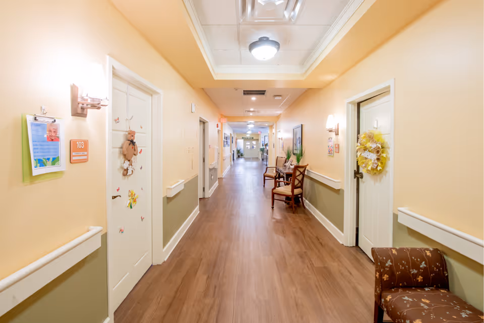 A well-lit hallway in a senior living facility with light yellow walls and wood flooring. Doors line both sides of the corridor, some decorated with wreaths and ornaments. There are chairs and small tables with plants along the right side of the hallway. The ceiling has recessed lighting and a central light fixture. An exit sign is visible at the far end of the hallway.