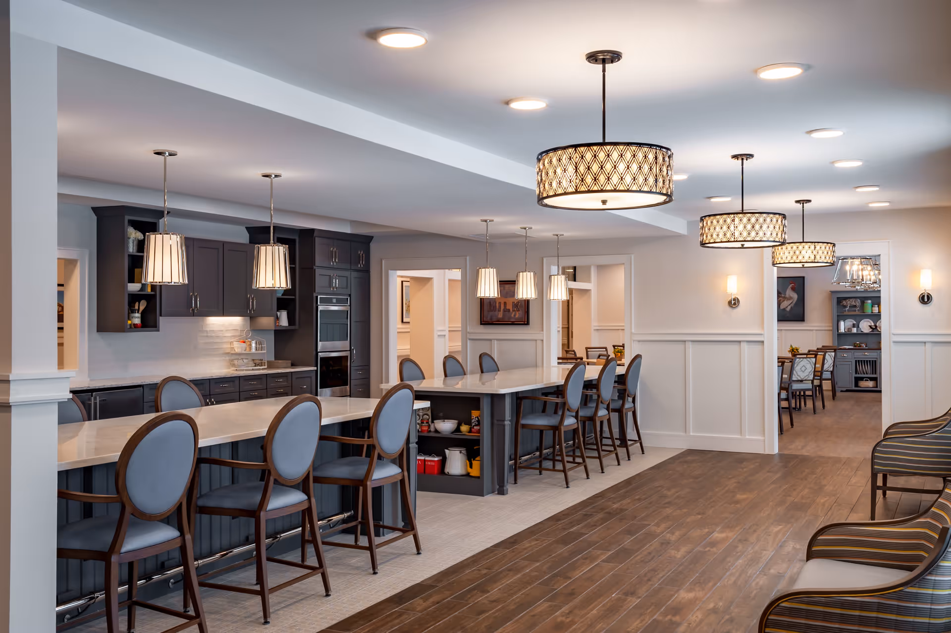A spacious, modern kitchen and dining area in a senior living facility with a long island counter surrounded by high-back chairs. The kitchen features dark cabinetry, built-in ovens, and pendant lighting. Adjacent to the kitchen is a hallway leading to a dining room with tables and chairs. The room has wood flooring, white walls with wainscoting, and multiple ceiling lights.