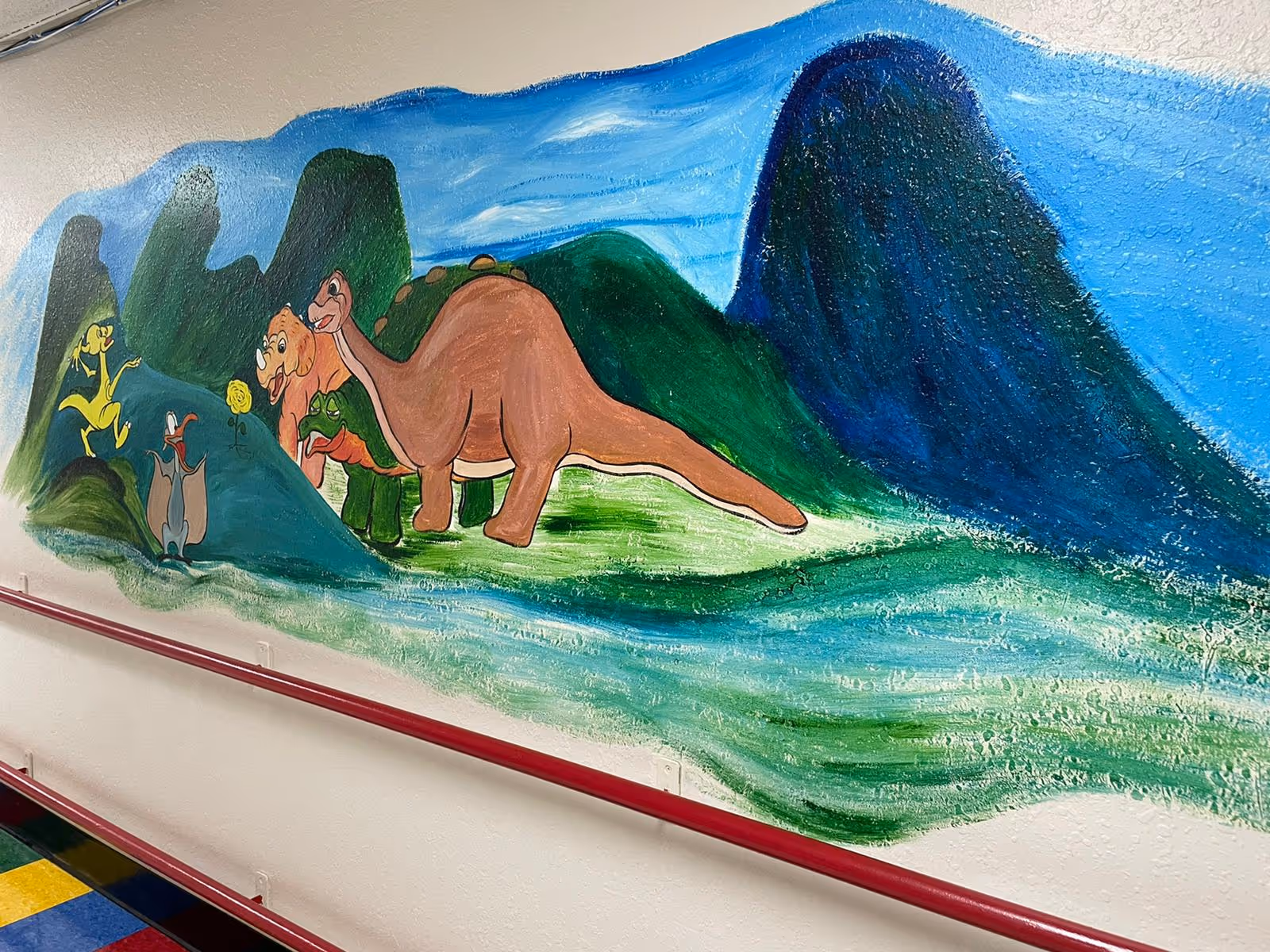 Colorful mural of cartoon dinosaurs and a green-blue landscape painted on an interior hallway wall with a red handrail and tiled floor.