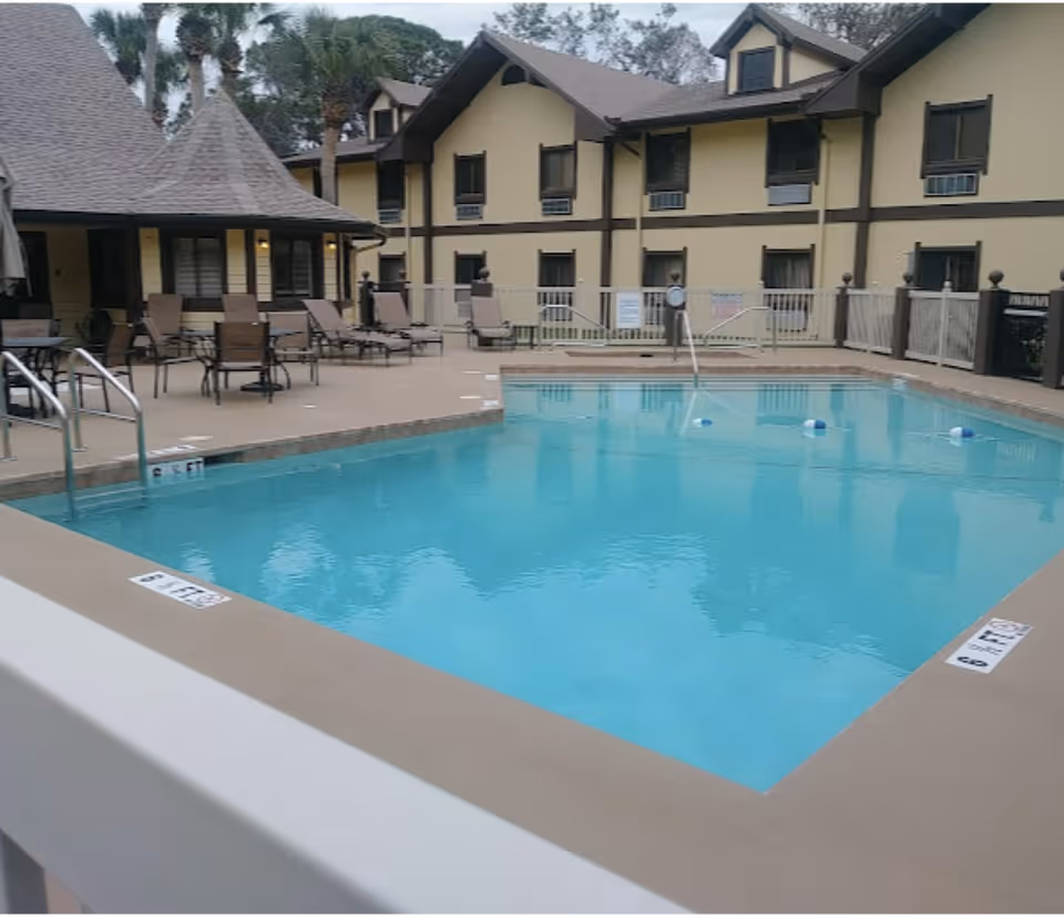 Outdoor swimming pool with clear blue water surrounded by a beige deck. Several lounge chairs and tables with chairs are arranged near the pool. In the background, there is a two-story building with a beige and brown exterior and multiple windows. Palm trees and other greenery are visible behind the building.