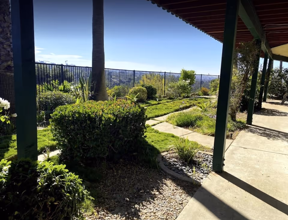 Covered patio walkway overlooking a landscaped garden with trimmed bushes, a palm tree, and a hillside view beyond a metal fence.