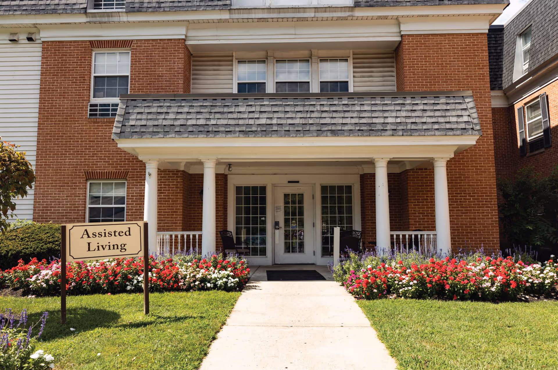 Exterior brick building with white columns, glass doors, and an assisted living sign by flowers.