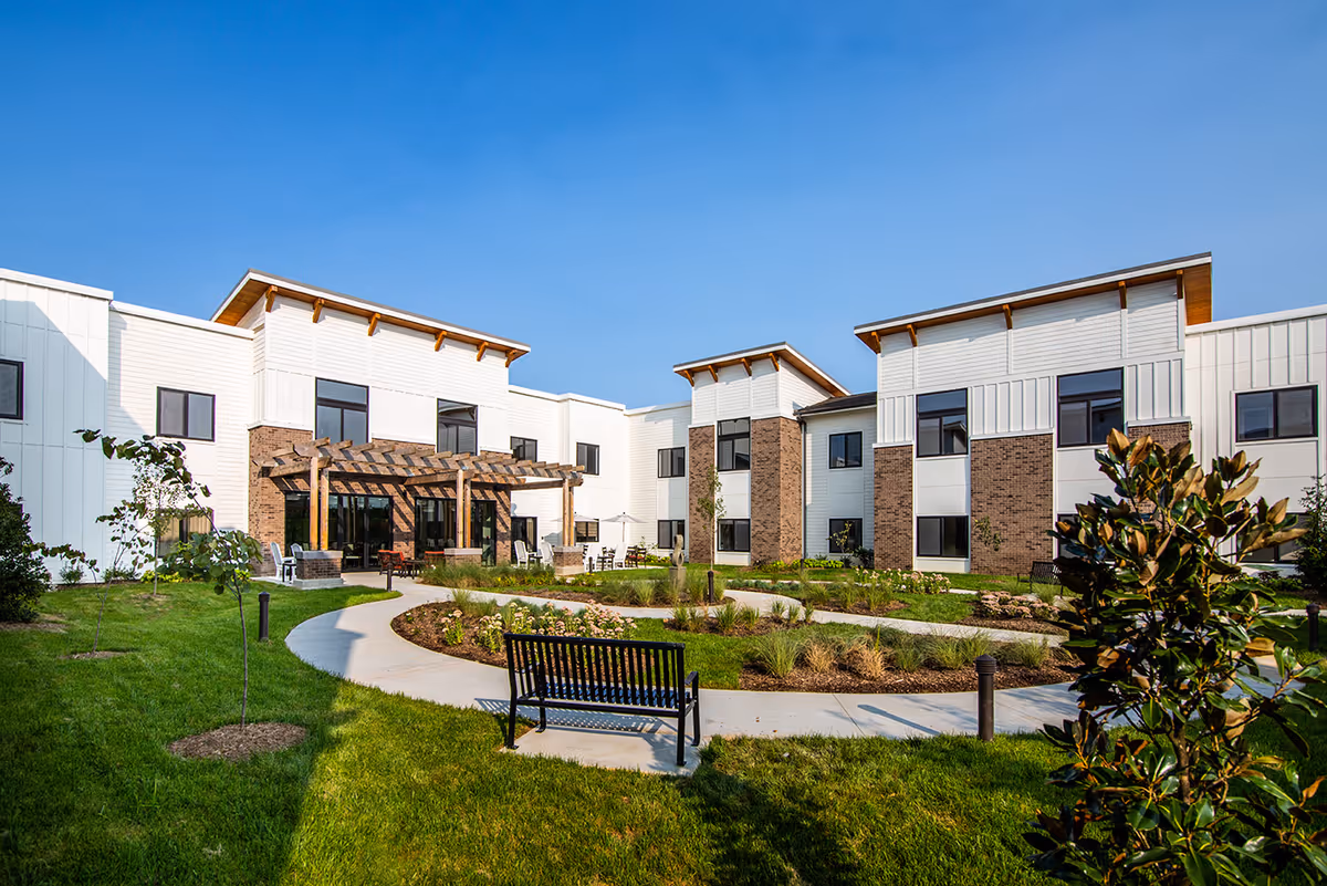Outdoor courtyard with pathways, benches, landscaping and a two-story senior living building with a pergola under a clear blue sky.
