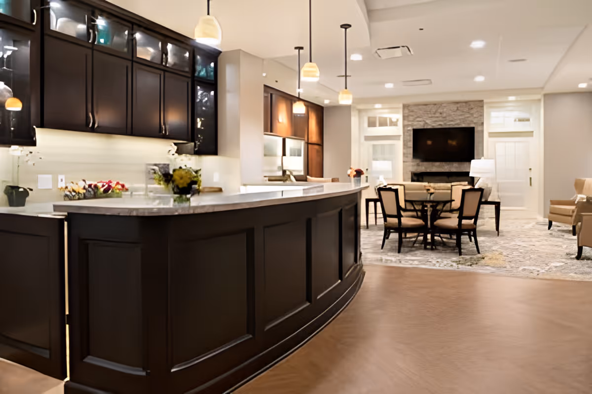 Interior view of a senior living facility showing a spacious kitchen area with dark wood cabinets and a curved marble countertop. Pendant lights hang above the counter. In the background, there is a dining area with a round table and chairs, a stone fireplace with a mounted TV above it, and comfortable seating including armchairs. The room is well-lit with recessed ceiling lights and has a warm, inviting atmosphere.