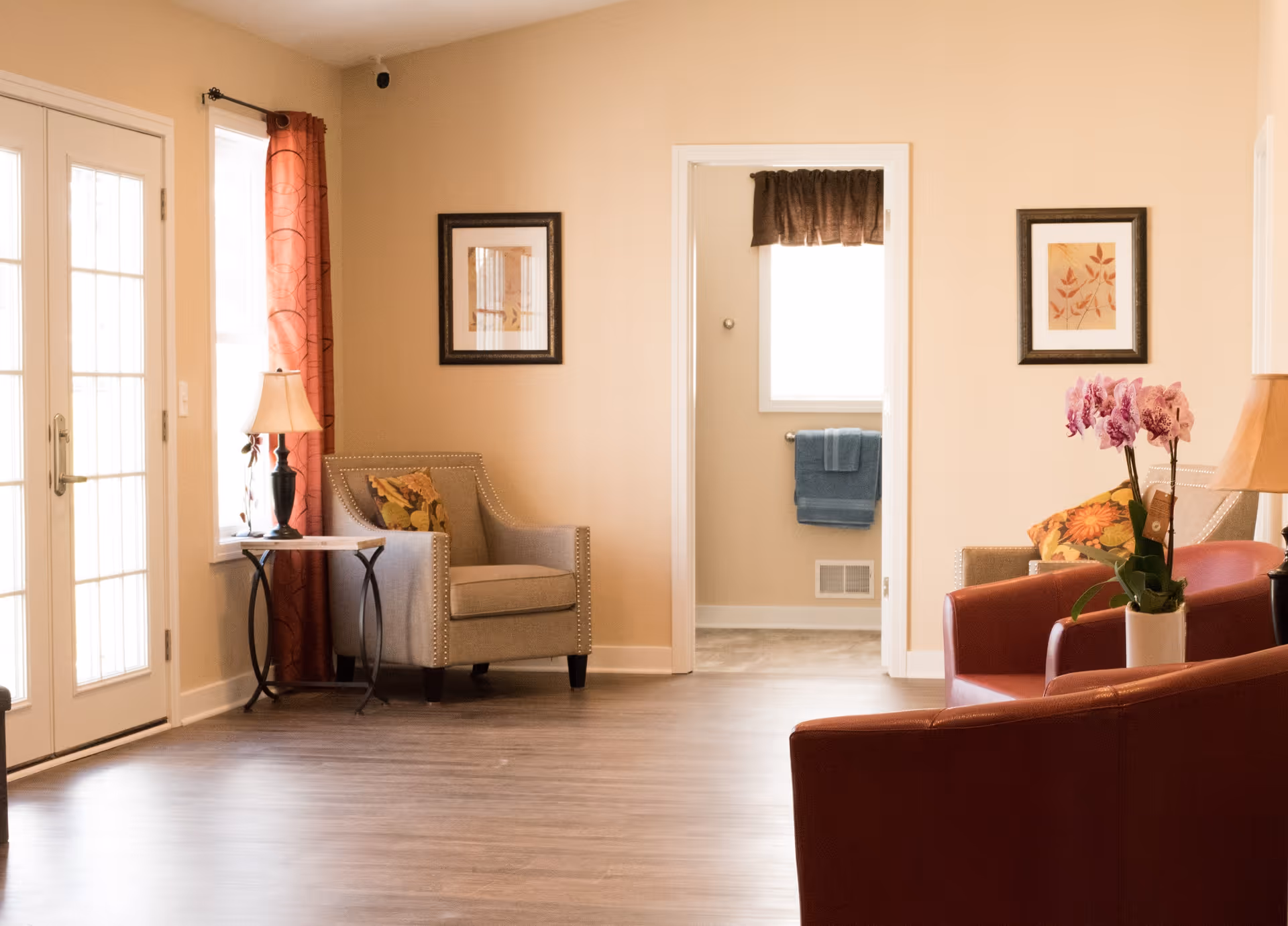 Bright assisted-living common room with armchairs, side tables, framed art, and a doorway leading to a bathroom.