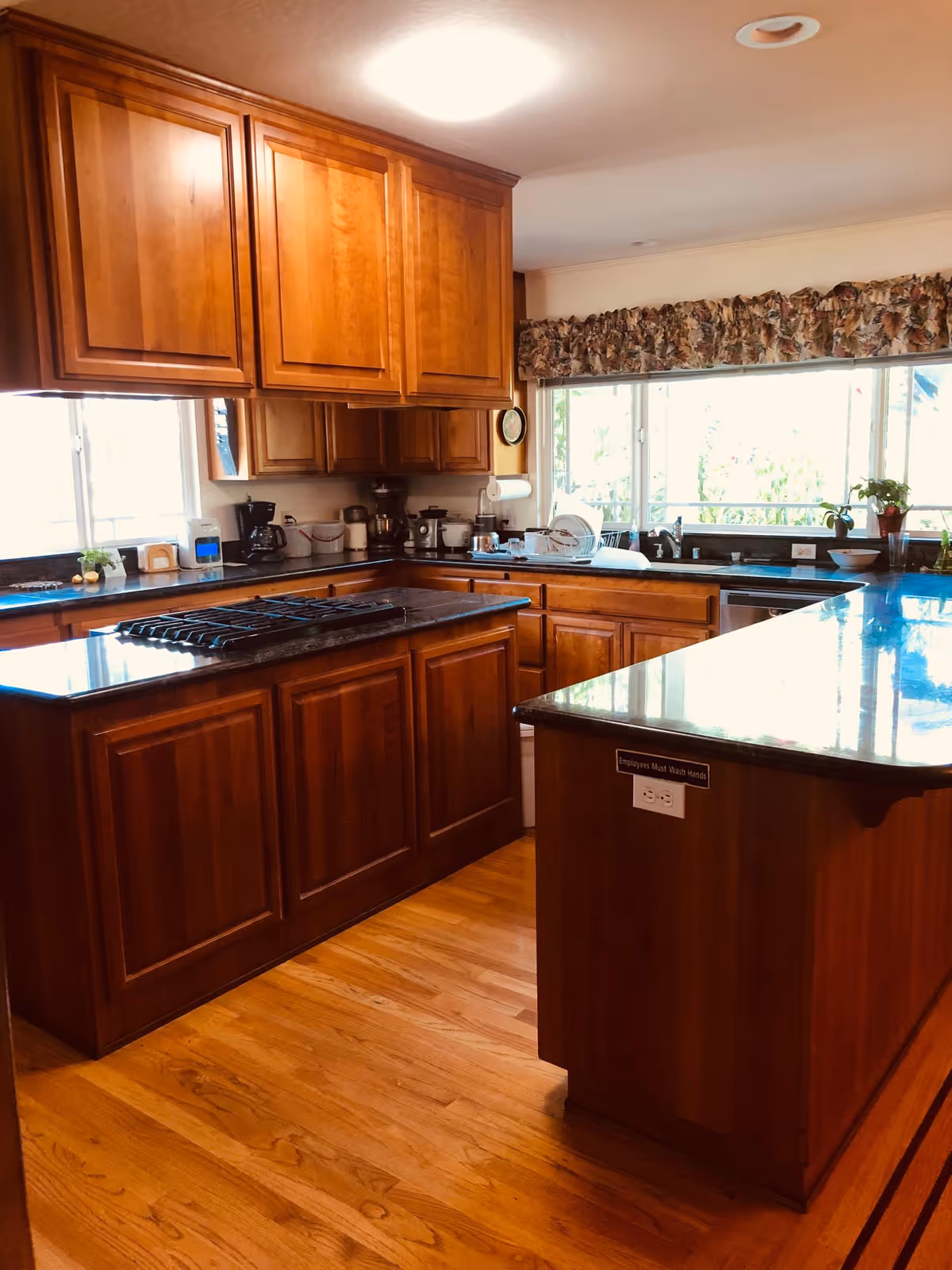 A kitchen with wooden cabinets and hardwood floors. There is a large island with a stovetop in the center and another counter with a sink near a window with floral valance curtains. Various kitchen appliances and utensils are on the counters.