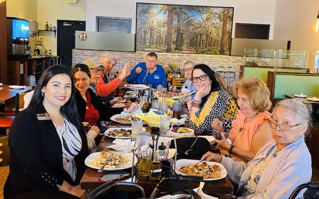 A group of elderly people and a staff member sitting around a dining table in a restaurant-style setting, enjoying a meal together. The table is filled with plates of food, glasses of water, and condiments. The background features a large painting of palm trees and a brick wall.