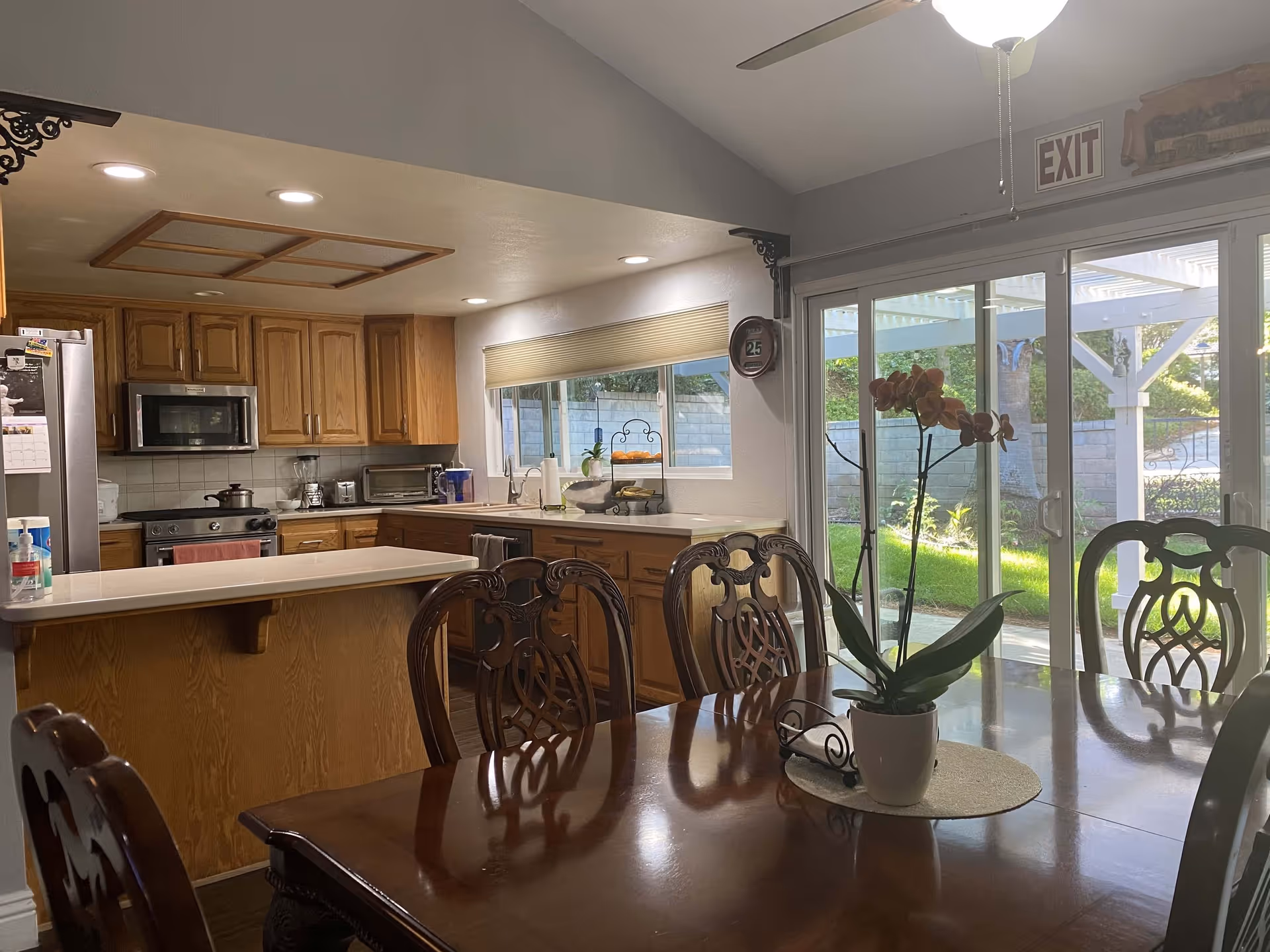 Interior view of a kitchen and dining area in a senior living facility. The kitchen features wooden cabinets, a stainless steel refrigerator, microwave, stove, and various small appliances on the countertops. The dining area has a polished wooden table with ornate chairs and a potted orchid centerpiece. Large sliding glass doors lead to an outdoor patio with greenery visible outside. An exit sign is mounted above the sliding doors.