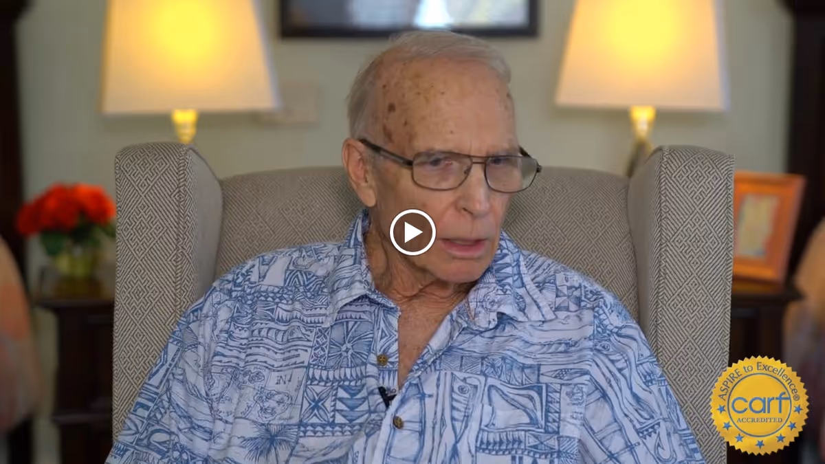 An elderly man wearing glasses and a patterned blue and white shirt is seated in a beige armchair with a geometric design. Behind him, there are two lit table lamps and a small table with a vase of red flowers and a framed picture.