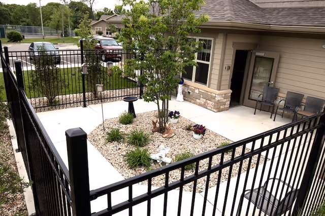 A small enclosed outdoor patio area with a black metal fence surrounding a concrete walkway and a central garden bed filled with rocks, plants, and a small tree. There are two chairs and a small table near the building entrance, and a parking lot with cars is visible in the background.