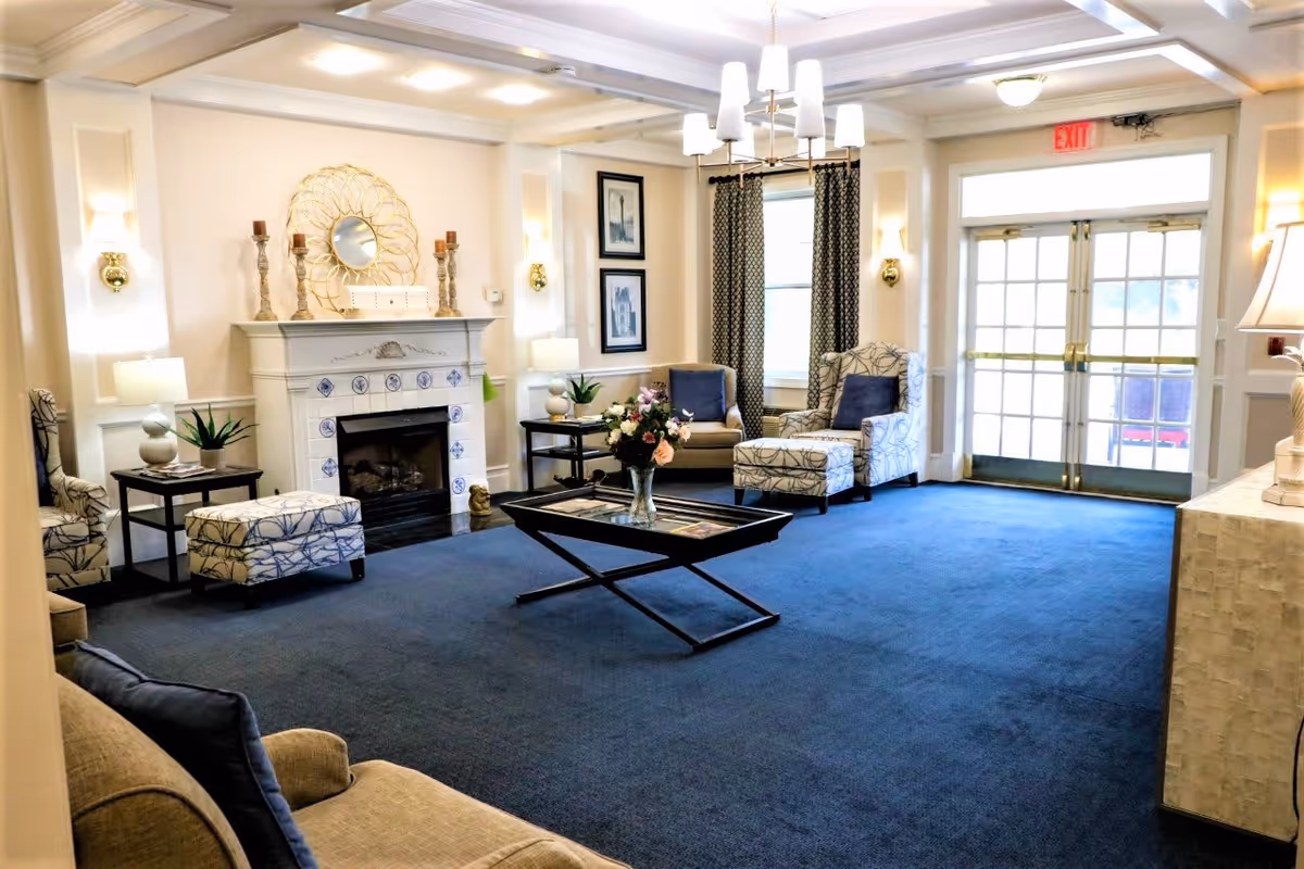 A bright and spacious living room area with blue carpet, beige walls, and a white fireplace decorated with candles and a round mirror. The room features patterned armchairs with blue cushions, a glass coffee table with a flower vase, side tables with lamps, framed artwork on the walls, and large windows with patterned curtains. Double glass doors with brass handles lead outside, and ceiling lights illuminate the space.