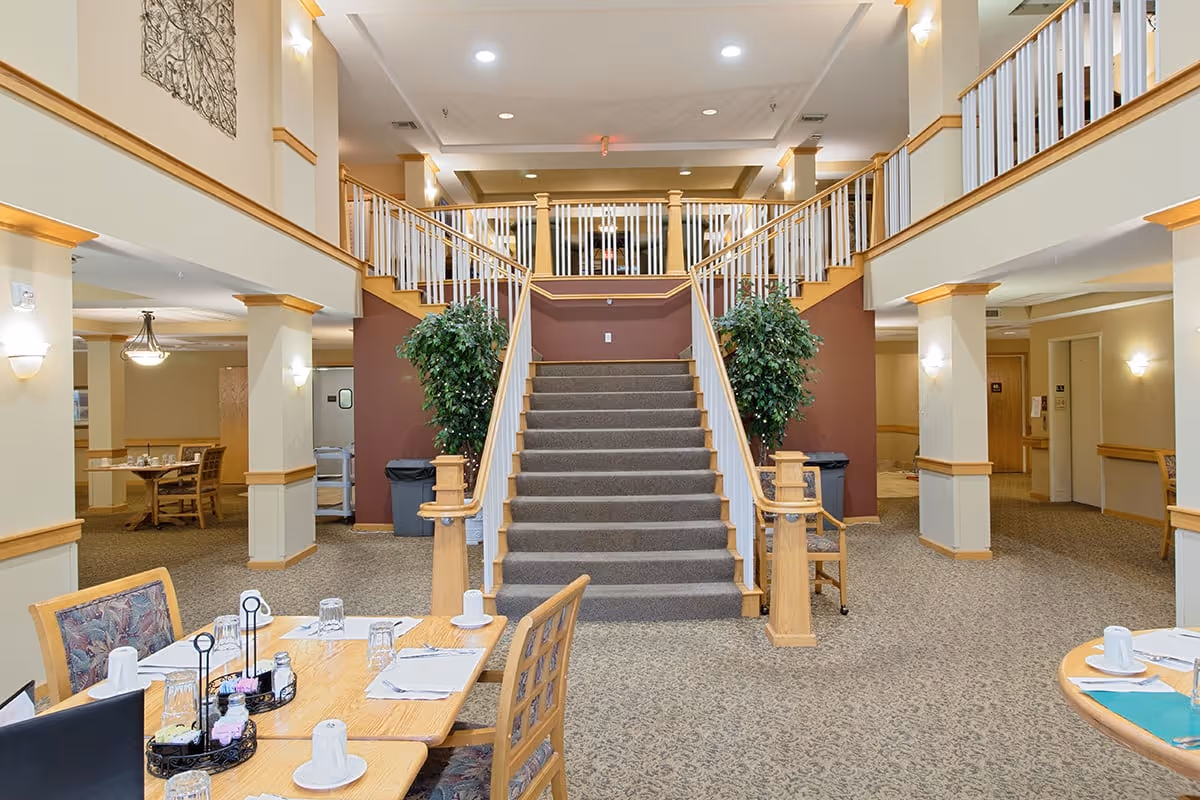 Interior view of Eagle Crest Assisted Living showing a dining area with tables set with cups, glasses, and condiments. In the background, there is a carpeted staircase with wooden railings leading to an upper floor, flanked by two large potted plants. The space is well-lit with wall sconces and ceiling lights, and there are additional seating areas visible to the left and right.