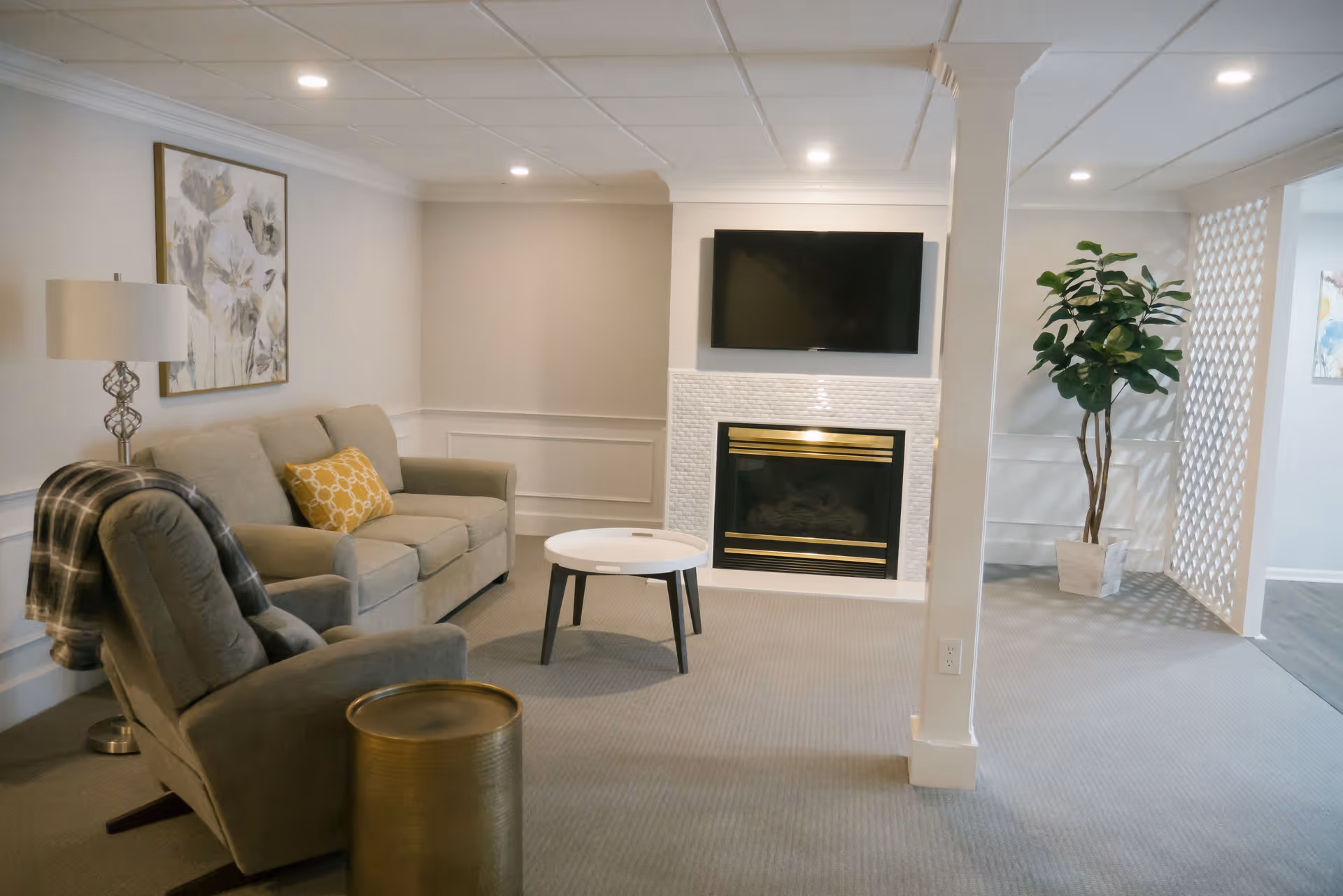 A cozy living room with a gray sofa and armchair, a round white coffee table, a floor lamp, a decorative painting on the wall, a fireplace with a mounted flat-screen TV above it, and a potted plant near a white lattice divider.