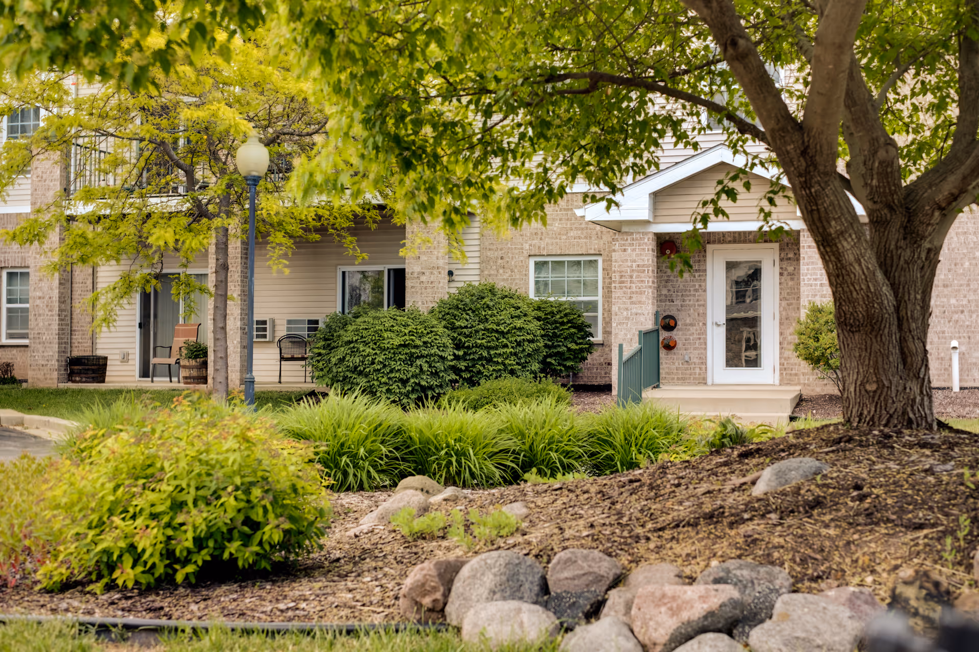 Exterior view of a senior living facility showing a landscaped garden with bushes, trees, and rocks in the foreground. The building has a brick facade with windows, a white door, and a small porch area with chairs. A streetlamp is also visible near the entrance.