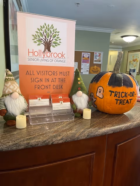 A reception desk at Hollybrook Senior Living of Orange decorated for Halloween with two gnome figurines, a large painted pumpkin with 'Trick-or-Treat' written on it, two small candles, and a sign instructing visitors to sign in at the front desk.