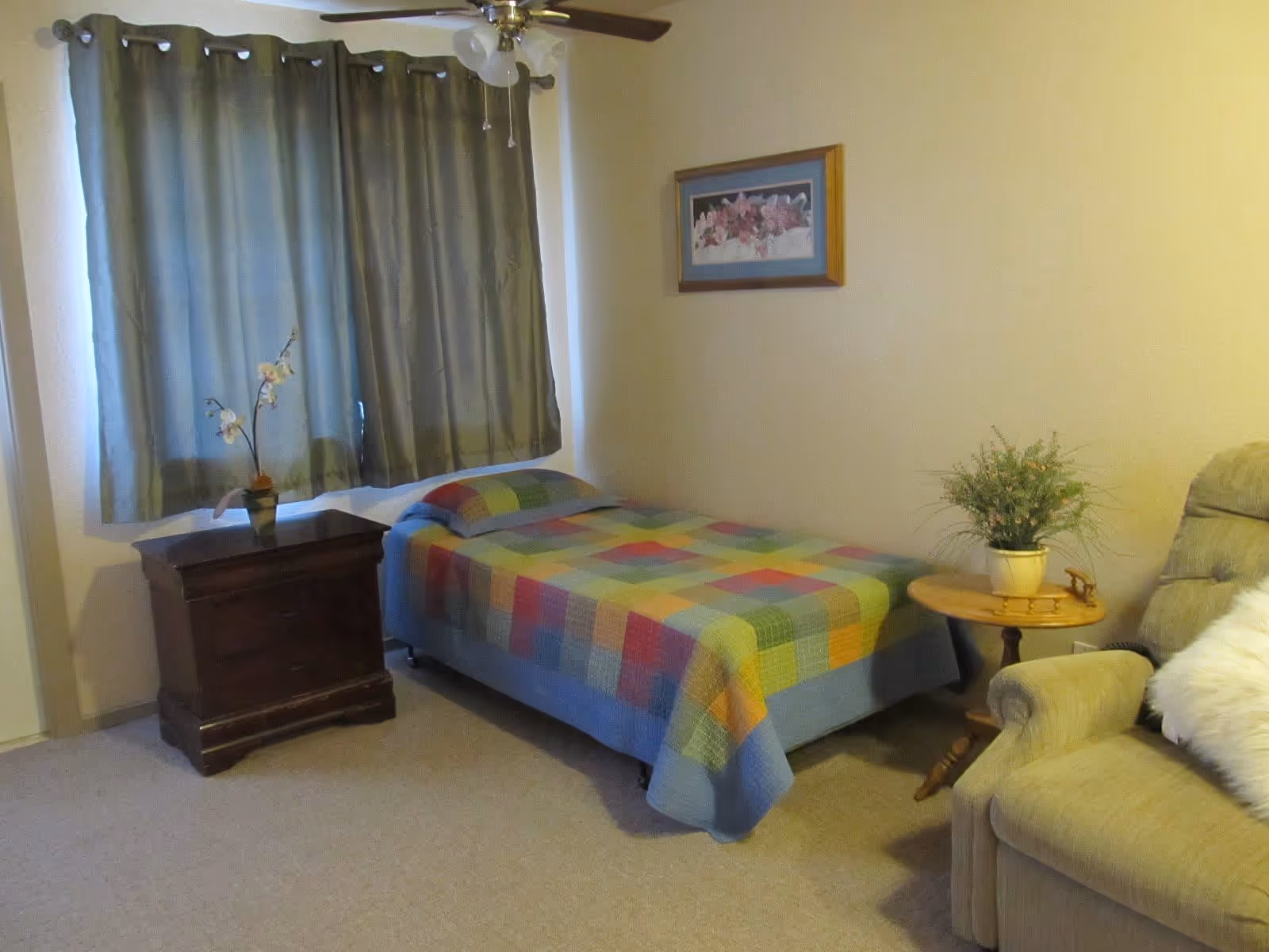 A small bedroom with a single bed covered in a colorful patchwork quilt. Next to the bed is a wooden nightstand with a small potted orchid on top. A window with green curtains is behind the nightstand. On the right side of the bed is a round wooden side table with a potted plant. A beige upholstered armchair with a white fluffy pillow is partially visible on the right. A framed floral picture hangs on the wall above the bed.