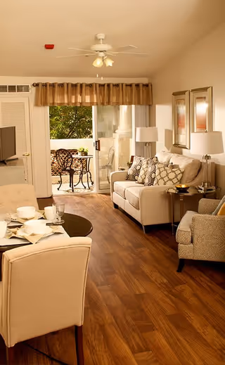 Well-lit living room with a sofa, armchairs, small dining table, wood floors, and a sliding glass door opening to a balcony with a bistro table.