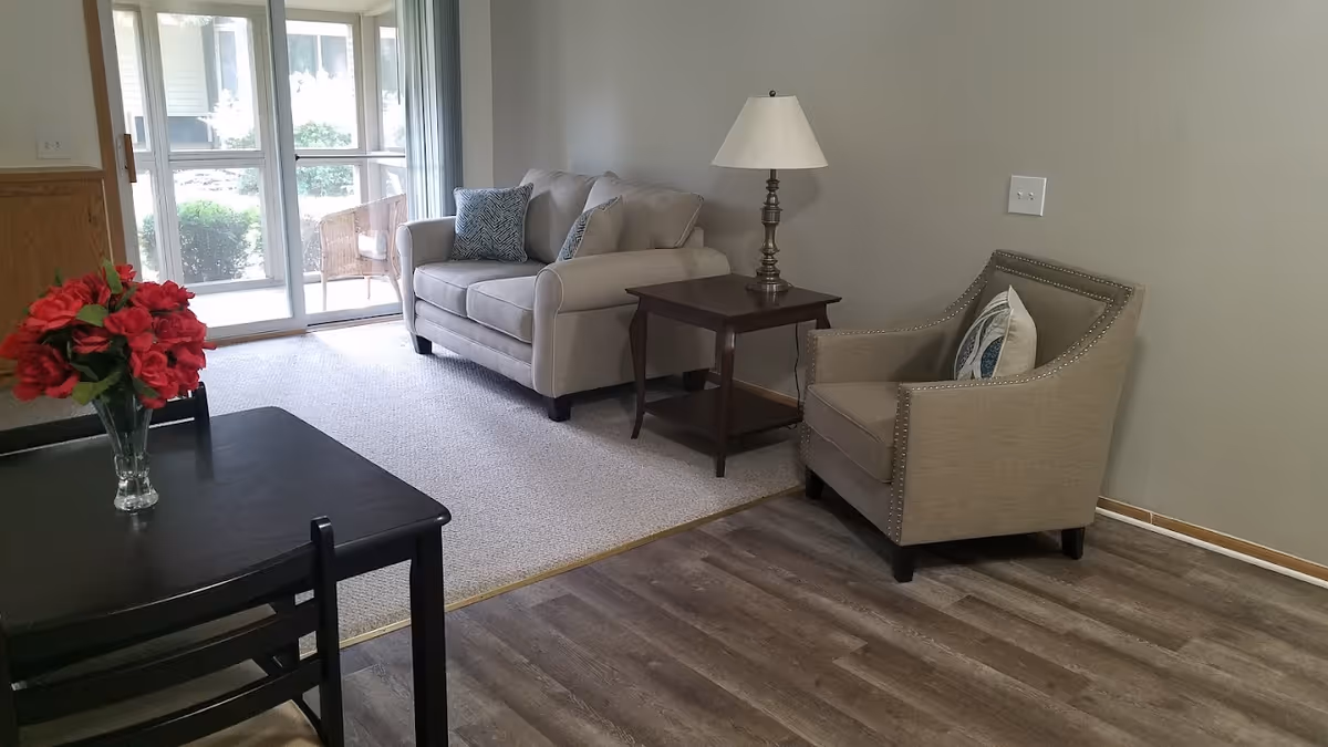 A cozy living room area with a beige loveseat and matching armchair, both with decorative pillows. A wooden side table with a lamp is placed between the loveseat and armchair. In the foreground, there is a dark wooden dining table with a vase of red flowers. The room has a mix of carpet and wood flooring, and a sliding glass door leads to an outdoor patio with chairs.