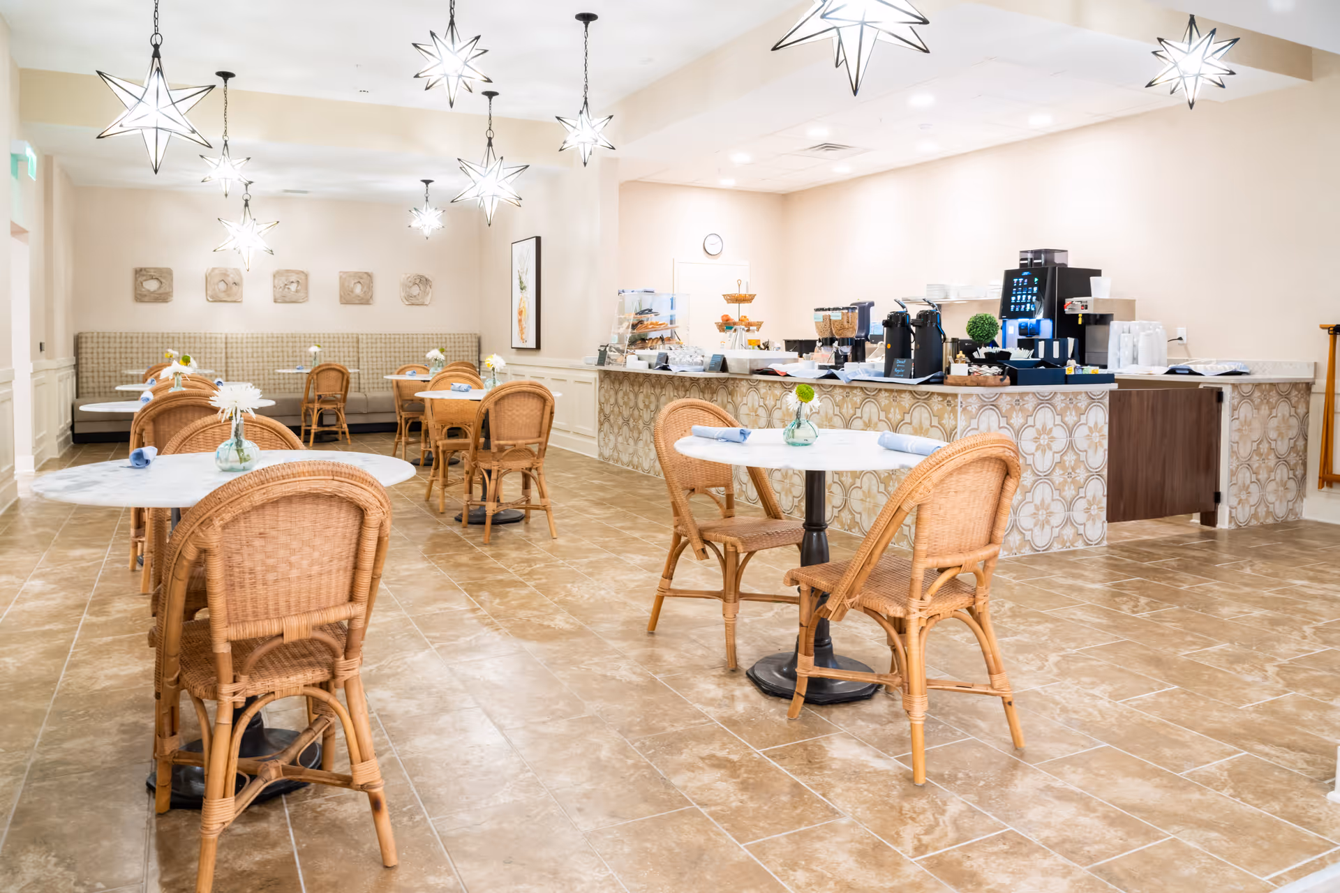 A bright dining area with several round tables topped with small vases of flowers and wicker chairs. The room features star-shaped pendant lights hanging from the ceiling and a counter area with coffee machines, cereal dispensers, and plates. The floor is tiled, and there is a cushioned bench along the back wall with decorative wall art above it.