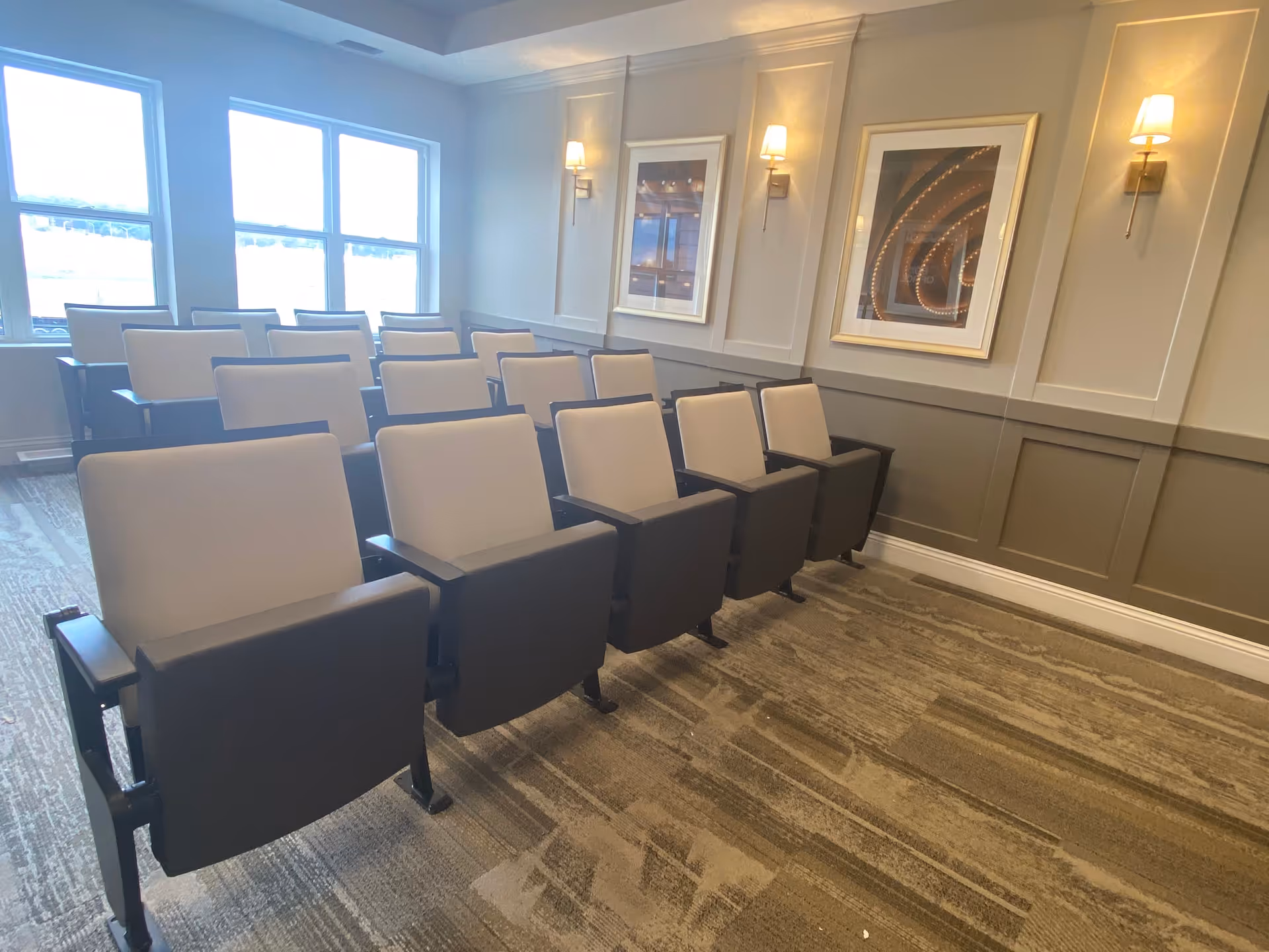 A small theater or presentation room with three rows of cushioned chairs facing forward. The room has large windows on one side letting in natural light, beige walls with decorative paneling, two framed pictures, and wall-mounted sconces providing warm lighting.