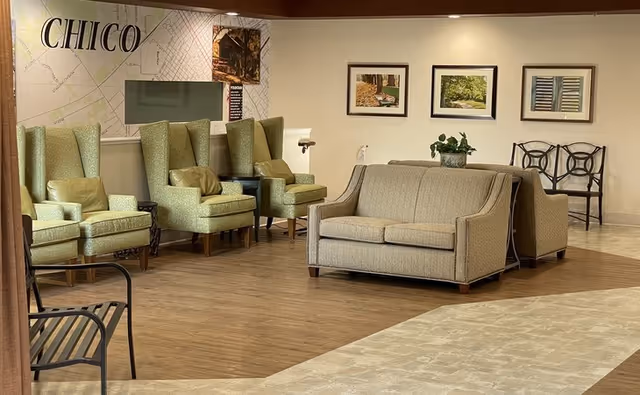 A comfortable seating area in a senior living facility featuring four green upholstered armchairs with cushions, a beige upholstered sofa, and a metal bench. The walls display framed pictures and a large map labeled 'CHICO'. The floor is a combination of wood and tile.
