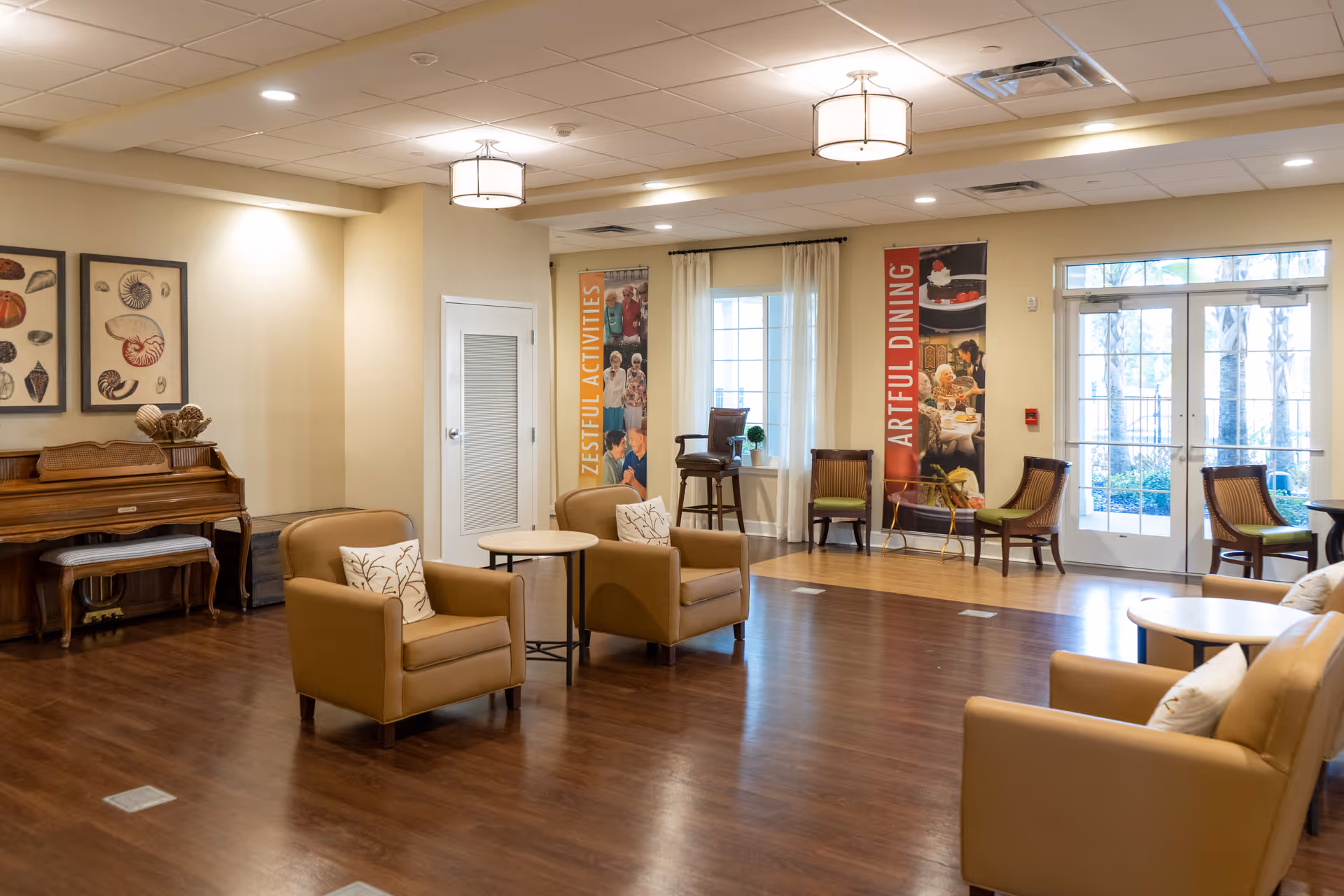 A spacious and well-lit common area with wooden flooring, beige walls, and ceiling lights. The room features several tan armchairs with decorative pillows arranged around small round tables. There is a wooden piano with a bench on the left side, and framed artwork above it. Two vertical banners hang on the far wall, one reading 'ZESTFUL ACTIVITIES' and the other 'ARTFUL DINING'. Large windows and glass doors allow natural light to enter the room, and additional chairs are placed near the windows.