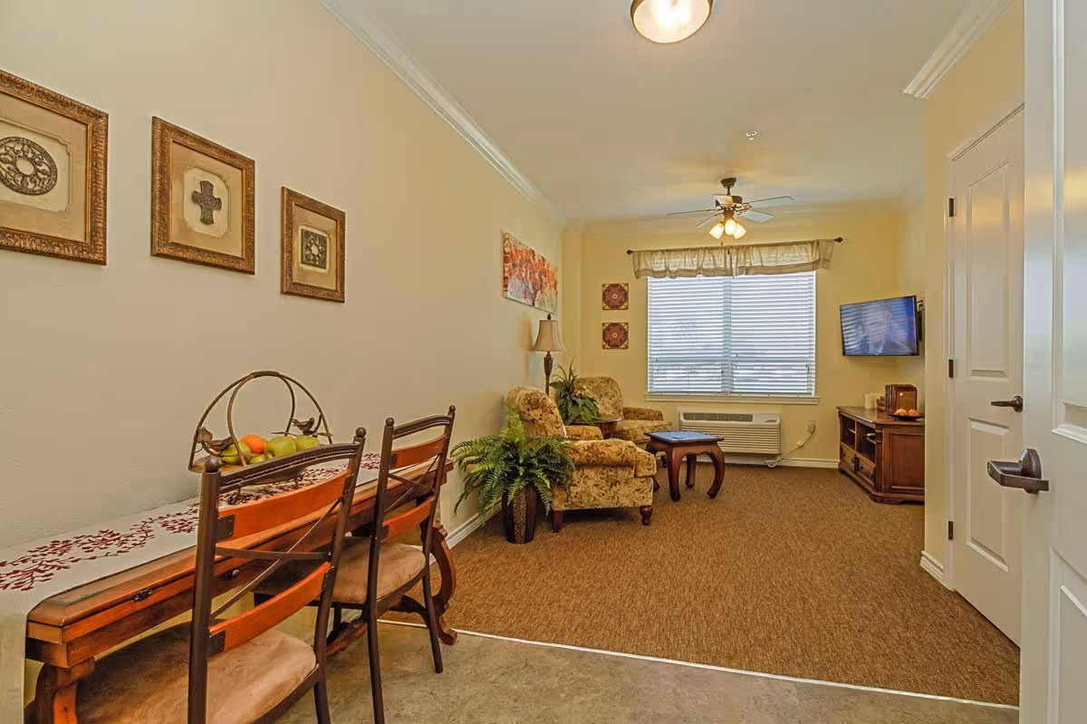 A cozy living room area in an assisted living facility featuring two floral upholstered armchairs, a wooden coffee table, a wall-mounted TV, and a window with blinds and a valance. To the left, there is a narrow wooden dining table with two chairs and a fruit basket on top. The walls are decorated with framed artwork and a table lamp is placed between the chairs.