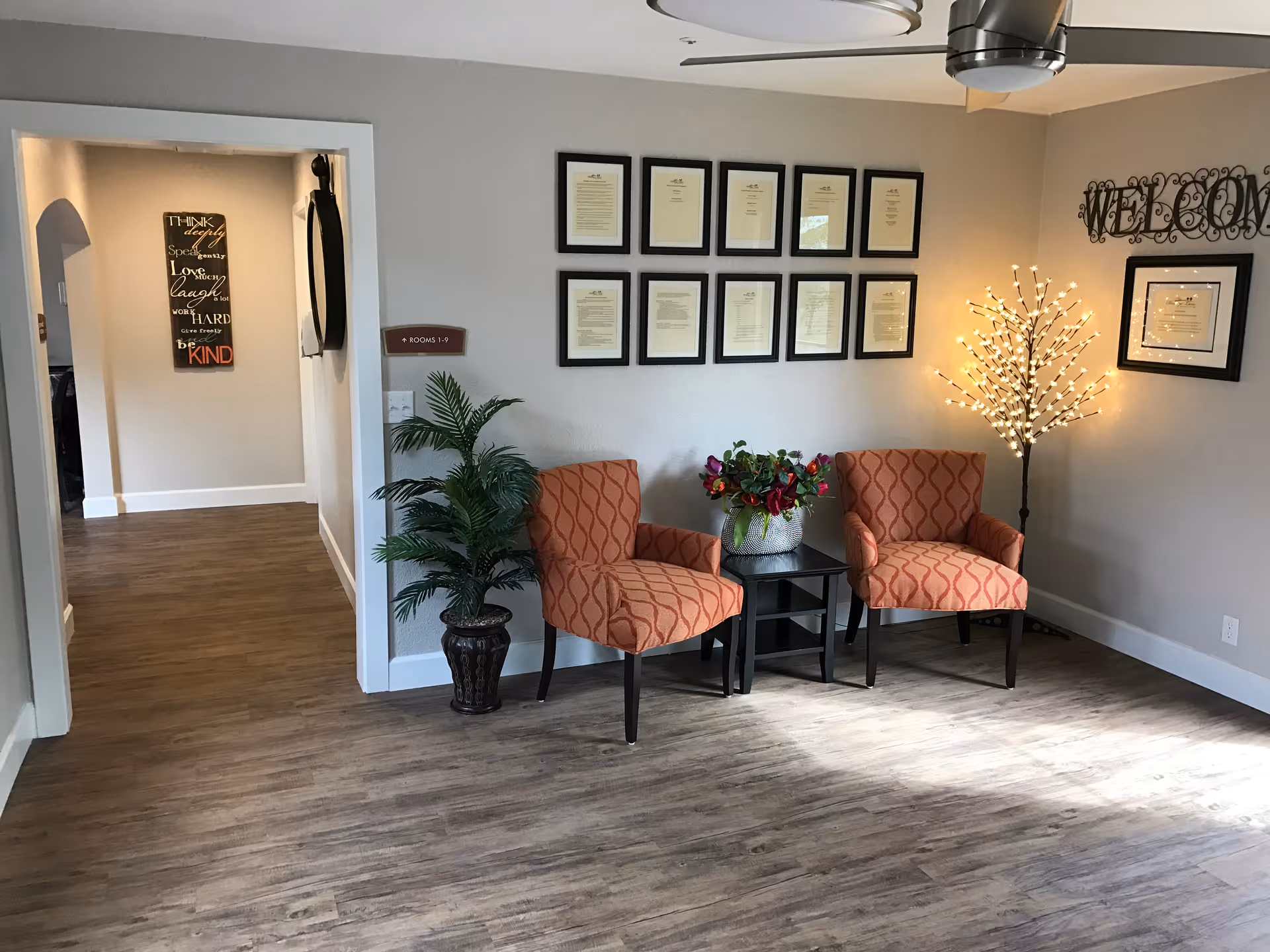 A cozy seating area in a hallway with two orange patterned armchairs separated by a small black table holding a flower arrangement. A decorative lit tree lamp stands beside the chairs. The wall behind features framed certificates and a metal 'WELCOME' sign. To the left, a hallway leads to other rooms with a sign indicating rooms 1-9 and a wall decoration with inspirational words.