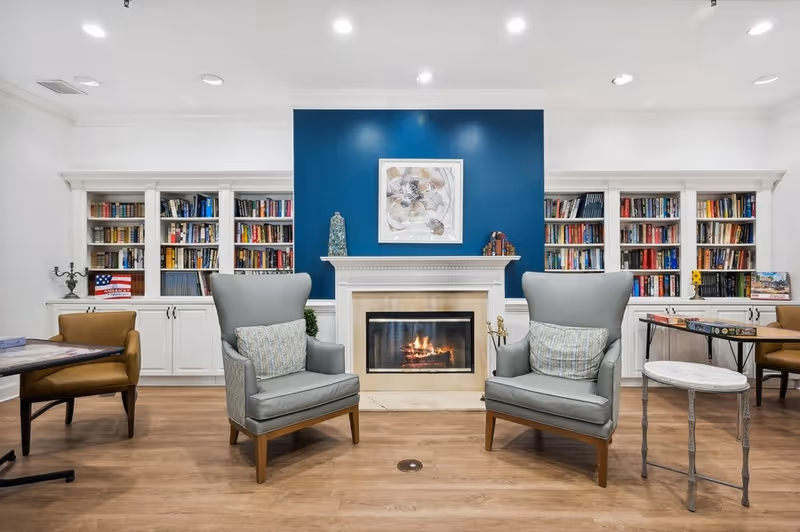 A cozy living room area with two gray armchairs facing a lit fireplace set in a white mantel with a blue accent wall behind it. The room has built-in white bookshelves filled with books on either side of the fireplace. There are additional chairs and tables on both sides, and the floor is wooden.
