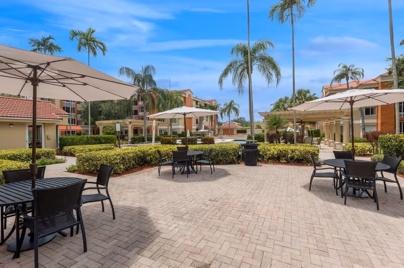 Outdoor patio area with several black tables and chairs under large white umbrellas, surrounded by neatly trimmed bushes and palm trees, with multi-story residential buildings in the background under a blue sky.