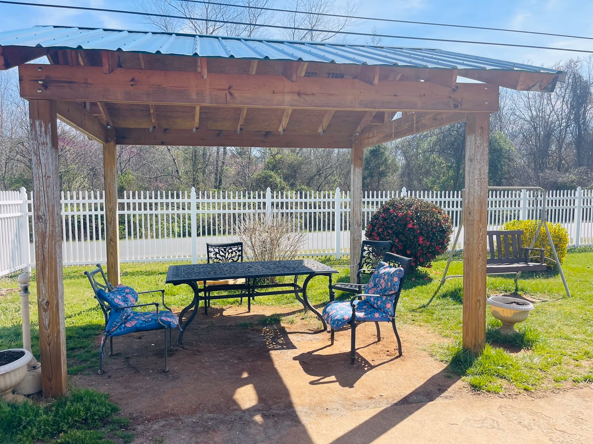Outdoor seating area with a wooden pergola providing shade over a metal table and four chairs with floral cushions. The area is surrounded by a white picket fence, green grass, bushes, and trees in the background. There is also a wooden swing bench and a small planter nearby.