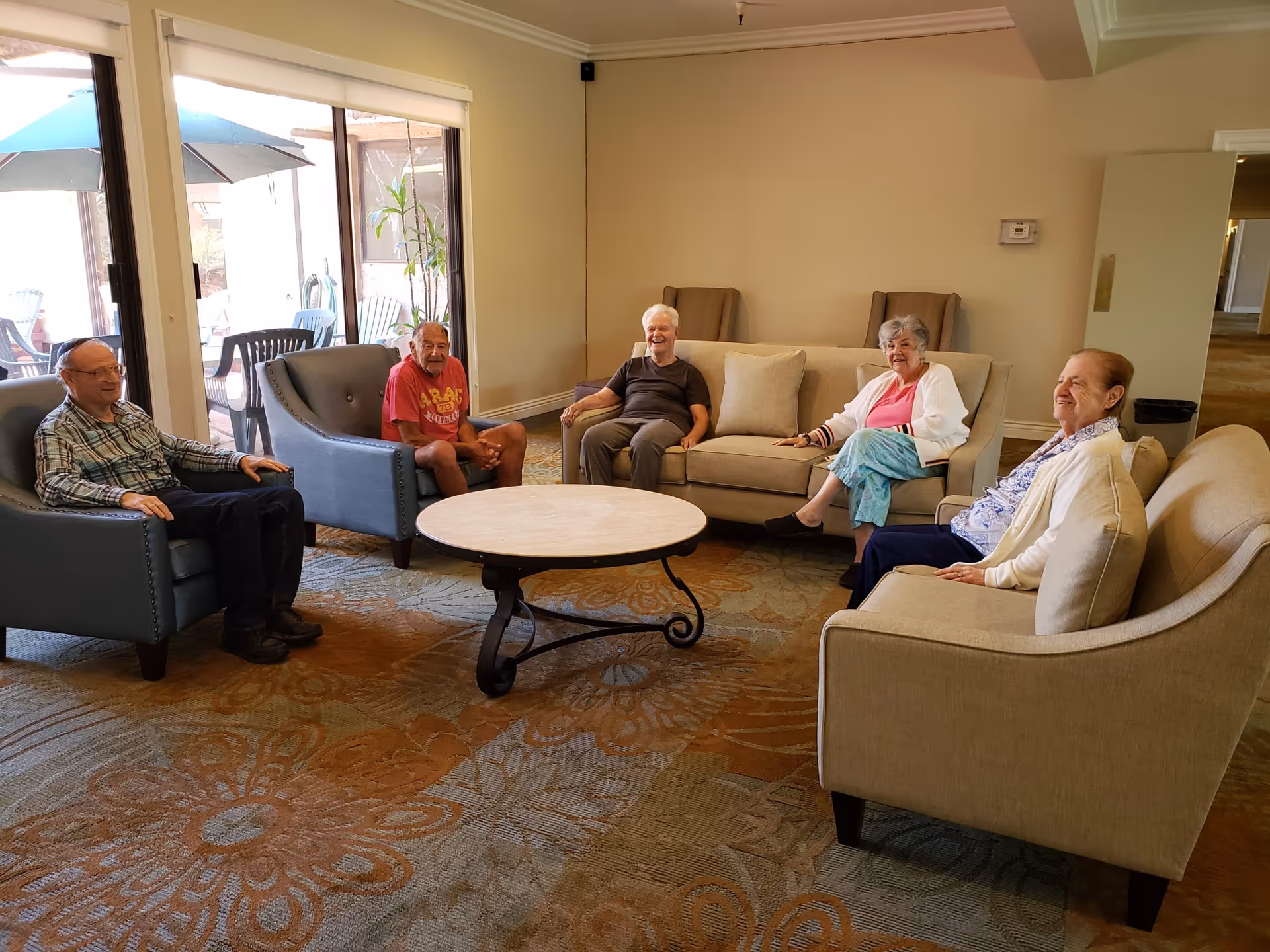 Five elderly people sitting in a living room area with beige and blue armchairs and sofas arranged around a round coffee table. Large windows show an outdoor patio with umbrellas and chairs. The room has a patterned carpet and neutral-colored walls.