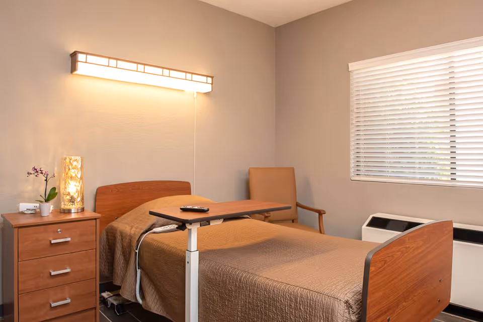 A simple bedroom in a senior living facility with a single bed covered in a brown quilt, a wooden bedside table with a small plant and lamp, a beige armchair, a window with white blinds, and a wall-mounted light above the bed.
