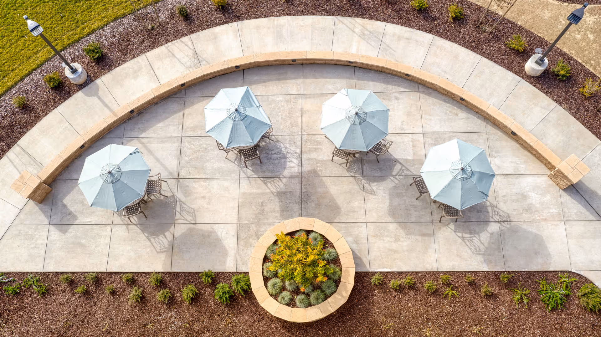 Aerial view of an outdoor patio area with four tables, each shaded by a light blue umbrella and surrounded by chairs. The patio is paved with concrete and bordered by a curved stone bench and landscaped garden beds with small plants and mulch. Two lamp posts are visible on either side of the patio.