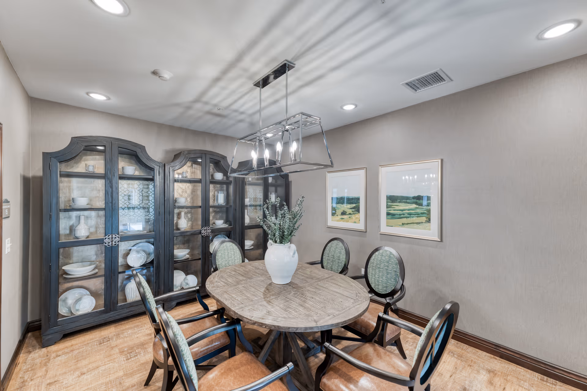 A formal dining room with a round wooden table surrounded by chairs, a large glass-front china cabinet, a modern chandelier, and framed landscape artwork on the wall.