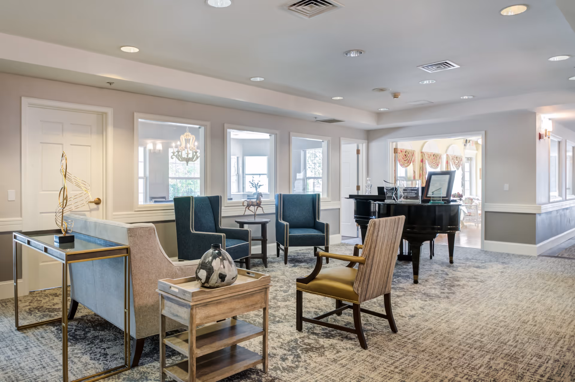 A spacious, well-lit common area with comfortable seating including two blue armchairs, a beige sofa, and a wooden chair with armrests. There is a small wooden side table with a decorative vase and a console table with a modern sculpture. In the background, a black grand piano is visible with framed items on top. The room has large windows and light-colored walls with carpeted flooring.