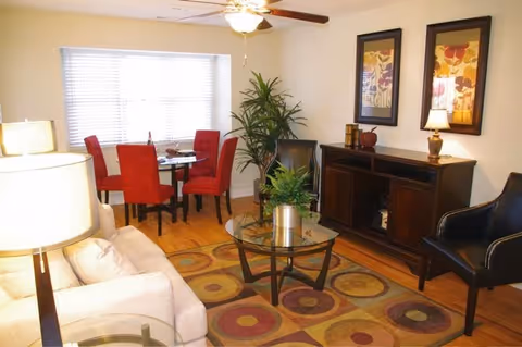 A cozy living room with a beige sofa, a glass coffee table with a plant, two black armchairs, a wooden sideboard with decorative items and two framed floral artworks above it. In the background, there is a dining area with a round glass table and four red upholstered chairs, next to a window with white blinds. The room has wooden flooring and a ceiling fan with a light.
