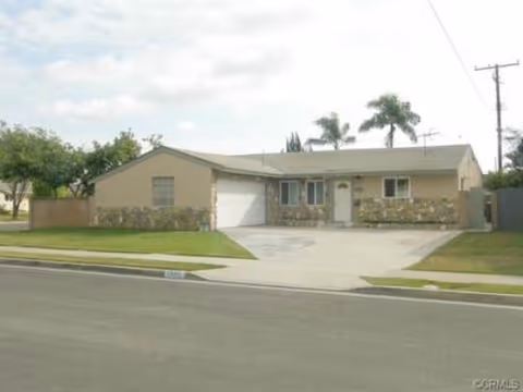 Single-story residential building with a stone and beige exterior, a garage, a driveway, and a front lawn with palm trees in the background under a partly cloudy sky.