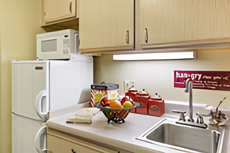 A small kitchen area with a white refrigerator, microwave on top, wooden cabinets, a countertop with a fruit basket, red canisters, and a sink with a faucet. There is an under-cabinet light illuminating the countertop.