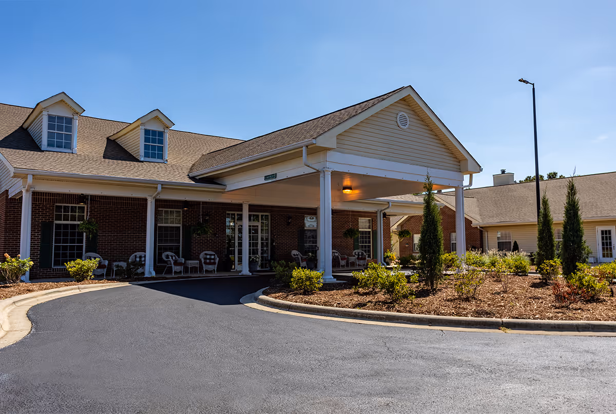 Front entrance of a single-story brick assisted living facility with a covered drop-off portico, porch chairs, and landscaped plantings.