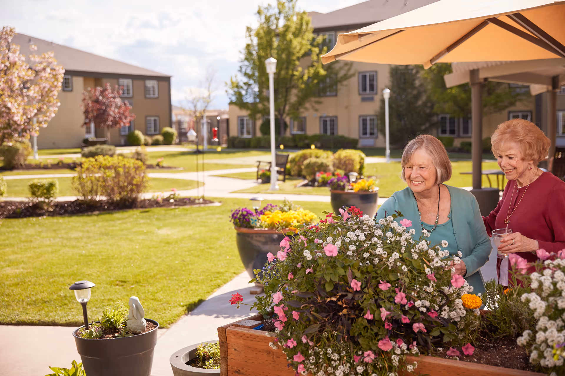 Two elderly women smiling and enjoying a sunny day while tending to colorful flowers in large planters outside in a garden area of a senior living facility. The background shows well-maintained lawns, trees, shrubs, and multi-story residential buildings under a partly cloudy sky.