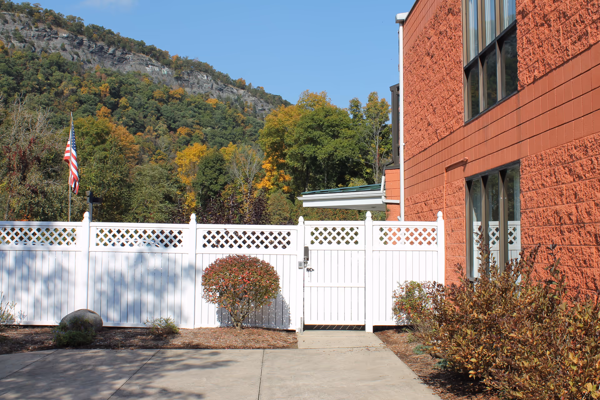 Outdoor view of a healthcare facility with a white fence and gate in front of a red brick building. There are bushes and trees around the area, with a mountain and blue sky in the background. An American flag is visible on the left side of the image.