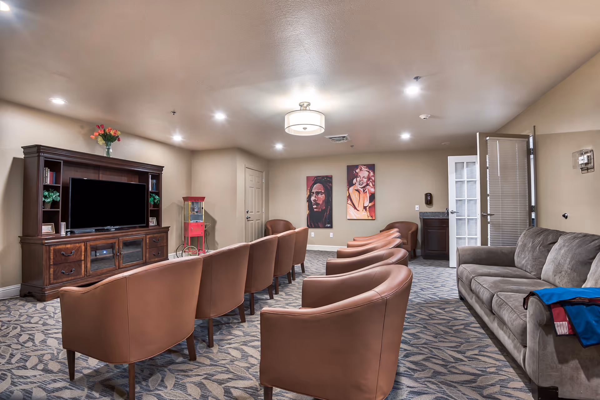 A senior living common TV room with rows of tan armchairs facing a wooden entertainment center, a gray sofa to the right, and artwork on the far wall.