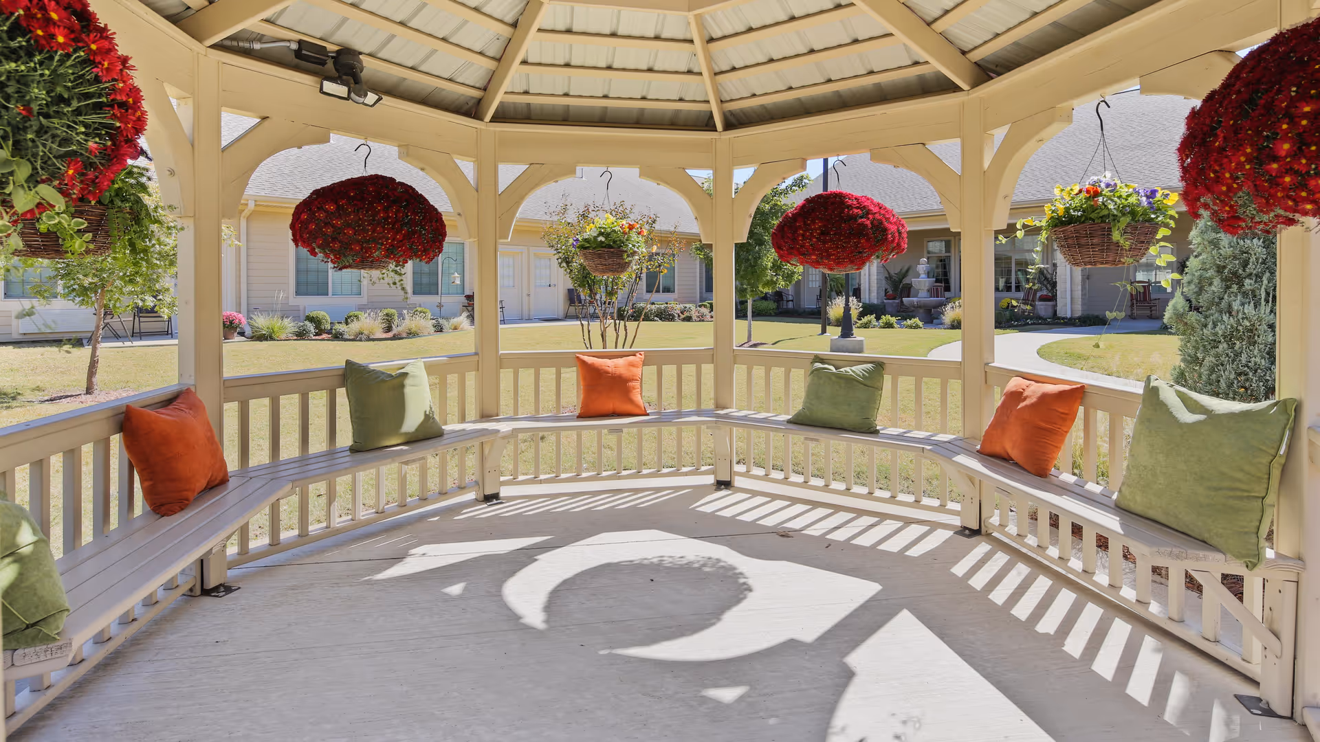 View from inside a gazebo with a circular bench adorned with green and orange cushions. Hanging baskets with red and multicolored flowers are suspended from the gazebo roof. The gazebo overlooks a well-maintained lawn and a building with beige siding and multiple windows.