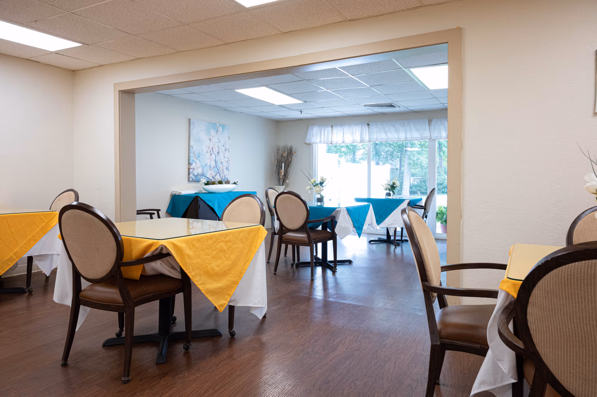 Interior view of a dining area in Parsons Residential Care Center with tables covered in white and yellow or blue tablecloths, surrounded by cushioned chairs. Large windows in the background let in natural light, and a painting and decorative plants are visible on the walls.