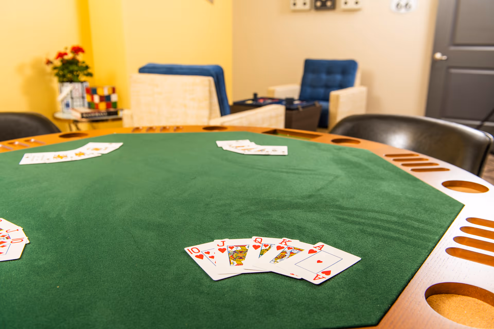 A green felt card table with playing cards arranged in a royal flush of hearts. In the background, there are two beige armchairs with blue cushions, a small table with a plant and books, and a closed gray door.