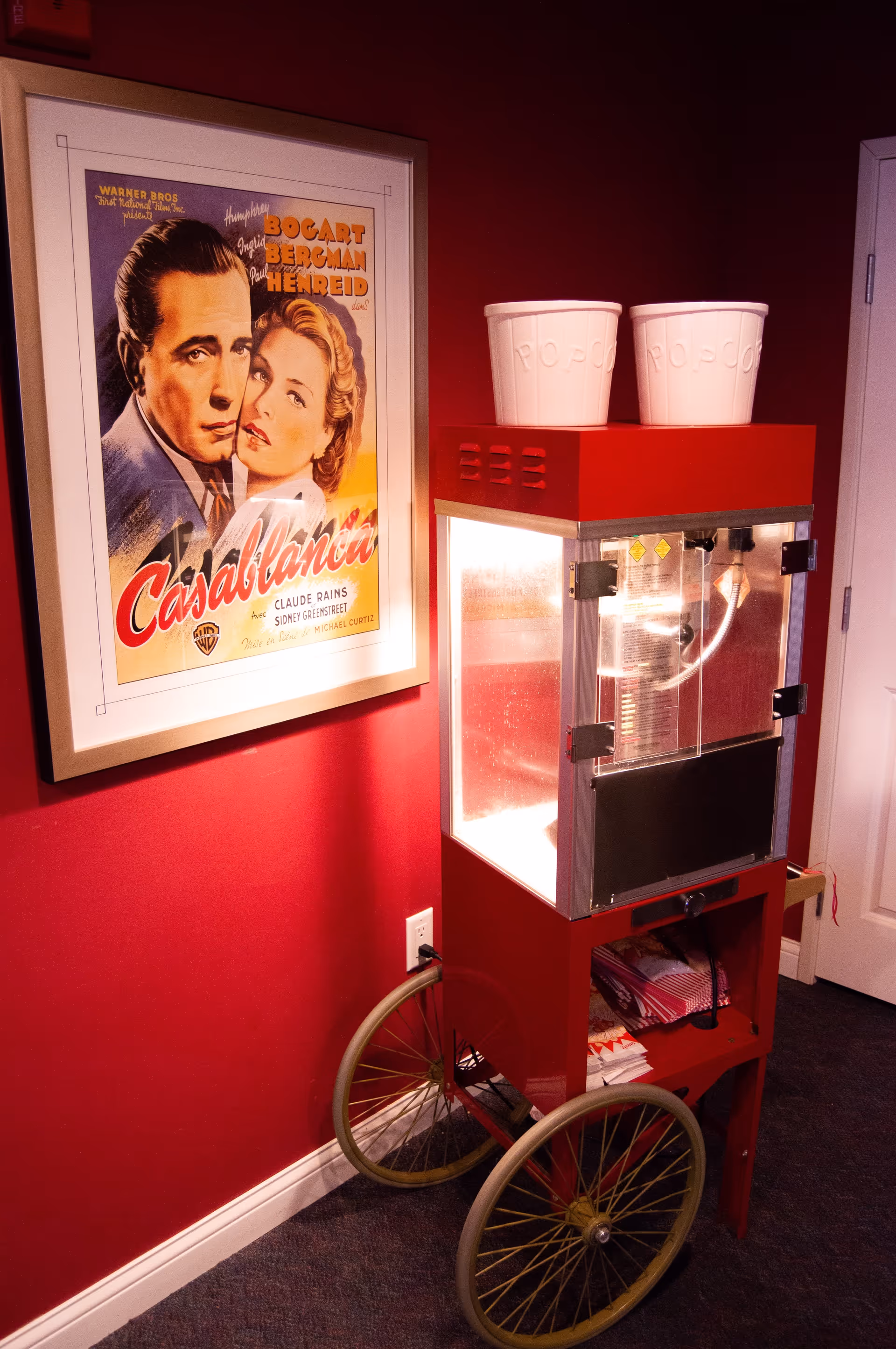 A vintage-style popcorn machine with two white popcorn buckets on top is placed against a red wall. Next to it, there is a framed Casablanca movie poster featuring Humphrey Bogart and Ingrid Bergman. The popcorn machine has large yellow wheels and is illuminated from within.