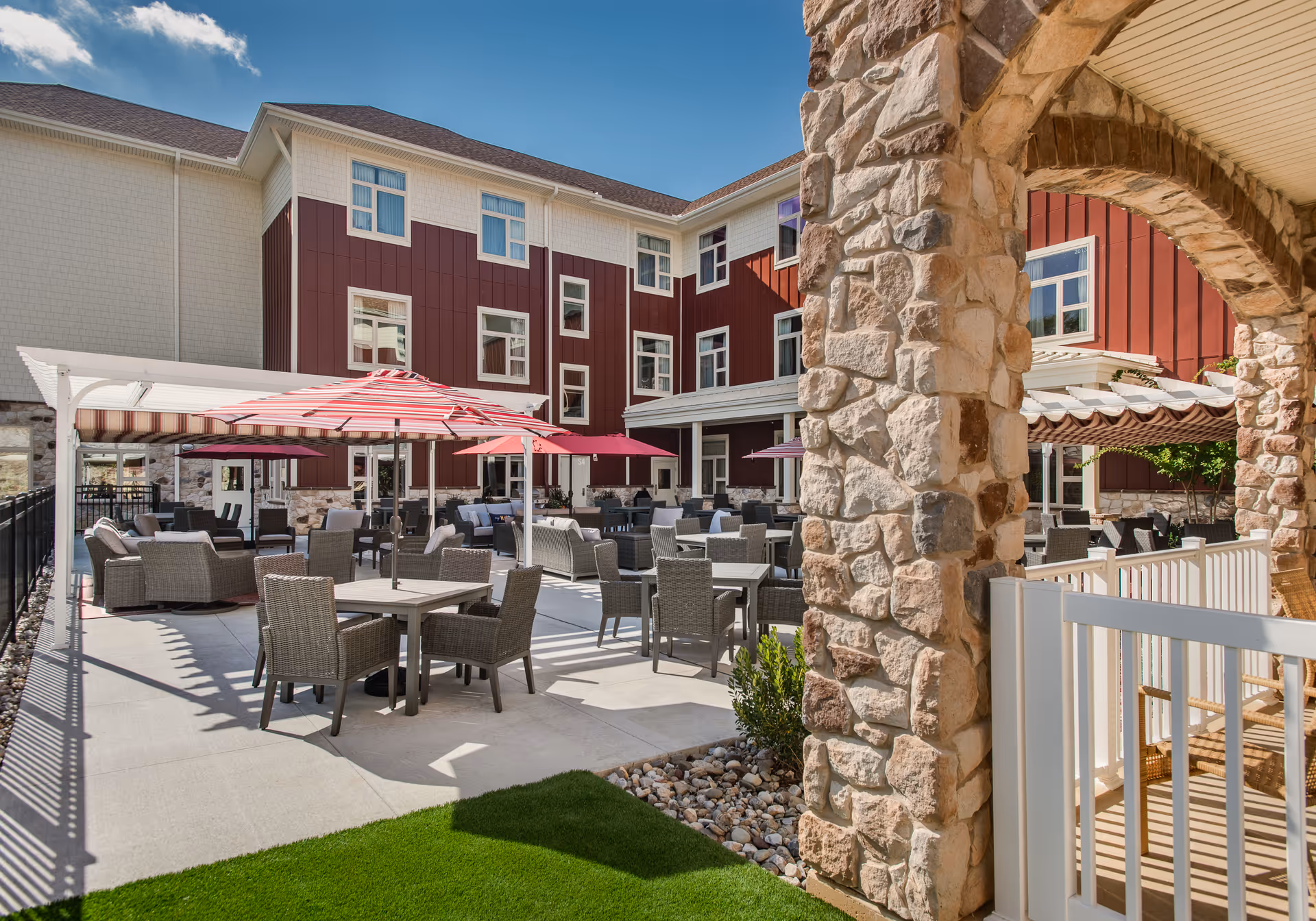 Outdoor patio area at Fieldstone at Chester Springs featuring multiple tables with chairs and umbrellas, surrounded by a three-story building with red and white exterior walls. Stone columns and a white railing are visible in the foreground under a clear blue sky.