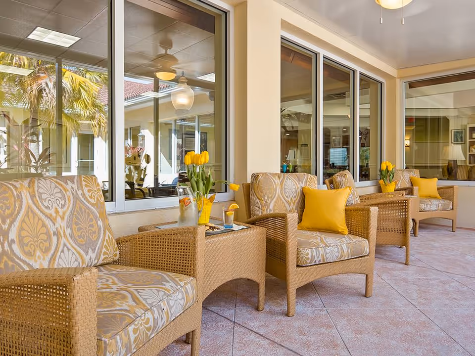 Covered outdoor seating area with wicker armchairs, patterned cushions, yellow throw pillows and vases of yellow tulips along a windowed wall.
